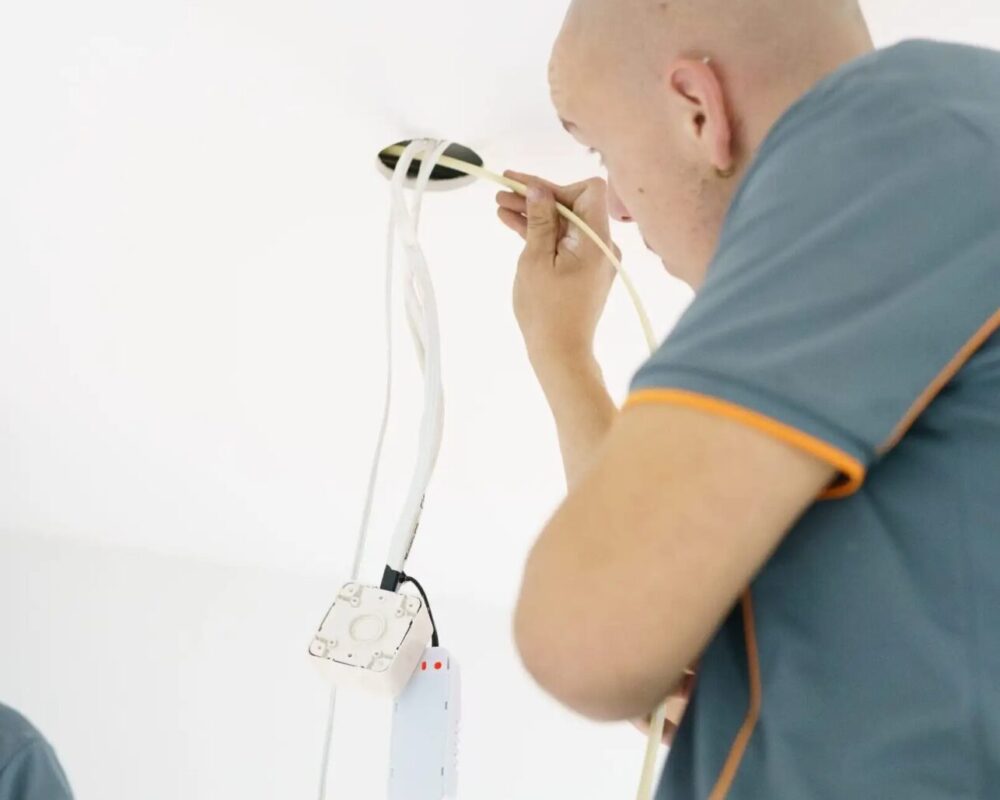 Electrician installing ceiling wiring and connecting a device while wearing a gray and orange uniform.