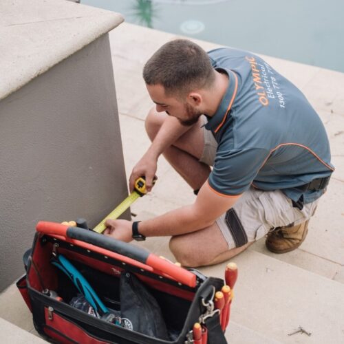 Technician kneeling and measuring next to a pool with a tape measure. Toolbag with various tools rests on the steps beside him.